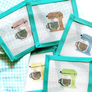 May include: A collection of five square needlepoint coasters. Each coaster features a stand mixer design with a coffee cup, in various colors including pink, orange, blue, yellow, and green. The coasters have a white background and are framed with teal fabric.