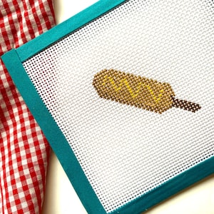 May include: A white canvas with a teal border, featuring a cross-stitch design of a yellow and brown hot dog.