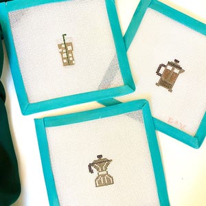 May include: Three square needlepoint coasters with turquoise frames. Each coaster features a different coffee-themed design: an iced coffee, a French press, and a stovetop espresso maker. The coasters are on a white surface.