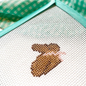May include: A white canvas with a brown and pink cross-stitch design of a bunny rabbit. The canvas is framed with a turquoise border.