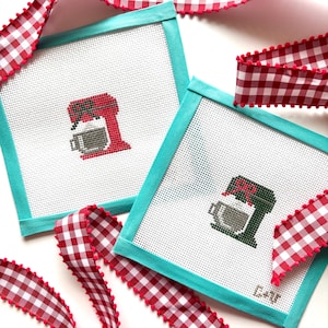May include: Two square needlepoint canvases with a teal border feature a stand mixer design in red and green. A red and white gingham ribbon with red pom-pom trim is draped around the canvases.