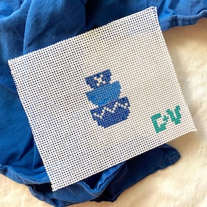 May include: A white needlepoint canvas with a blue geometric design and the letters "GV" in teal. The canvas is placed on a blue fabric, creating a contrast. The design features stacked shapes in varying shades of blue.