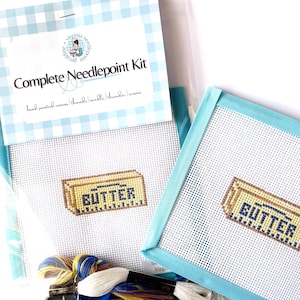 May include: A complete needlepoint kit featuring a hand-painted canvas with a butter stick design, including threads, a needle, a threader, and scissors. The packaging displays the text Complete Needlepoint Kit and Virginia Needlepoint.