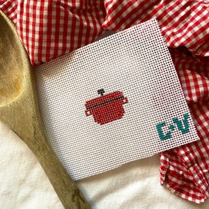 May include: A white canvas with a red pot and the letters "CV" in teal blue. The canvas is on top of a red and white checkered fabric.