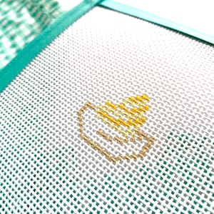 May include: Close-up of a white Aida cloth with a teal border. A small yellow and gold cross-stitch design is centered on the cloth.