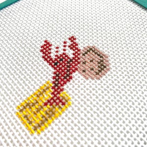 May include: A red and yellow cross-stitch design of a lobster with a brown and pink shell. The design is on a white canvas with a teal border.