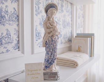 May include: A white and blue ceramic statue of a woman holding a child, featuring a floral pattern on the dress and a halo-like headpiece. A square sign next to the statue displays the text: Virgenita de la salud lirio, 22 cm.