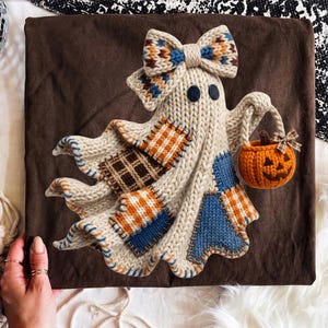 May include: A dark brown t-shirt featuring a graphic of a cream-colored knitted ghost with a matching bow. The ghost has dark circular eyes, patches in orange and blue plaid, and carries a small orange knitted pumpkin bucket with a brown bow.