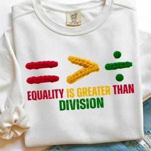 May include: White sweatshirt with the slogan "EQUALITY IS GREATER THAN DIVISION". The design includes red equal signs, a yellow greater-than symbol, and a green division symbol. It has a Comfort Colors label.