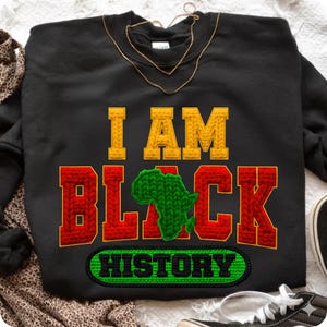 May include: Black sweatshirt with the text "I AM BLACK HISTORY" in yellow, red, and green. The word "BLACK" features a green Africa map graphic. A gold necklace is draped across the top of the sweatshirt.