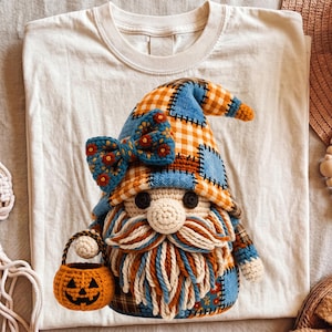 Könnte beinhalten: Ein cremefarbenes T-Shirt mit einer Grafik eines gehäkelten Gnoms, der einen blau-orange karierten Hut, eine große blaue Blumen-Schleife, einen dicken Bart aus cremefarbener und blauer Wolle sowie einen kleinen orangefarbenen Kürbiseimer trägt.