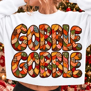 May include: A white long-sleeve sweatshirt featuring the text GOBBLE GOBBLE in large block letters. The letters are filled with a colorful pattern of overlapping leaf shapes in various autumn-inspired fabrics, including plaid, polka dots, and solids.