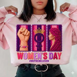 May include: Pink sweatshirt with a graphic design for Women's Day. The design includes a knitted fist, a female symbol with a crown, and a woman's profile. The text "WOMEN'S DAY" and "#INSPIREINCLUSION" are also featured.