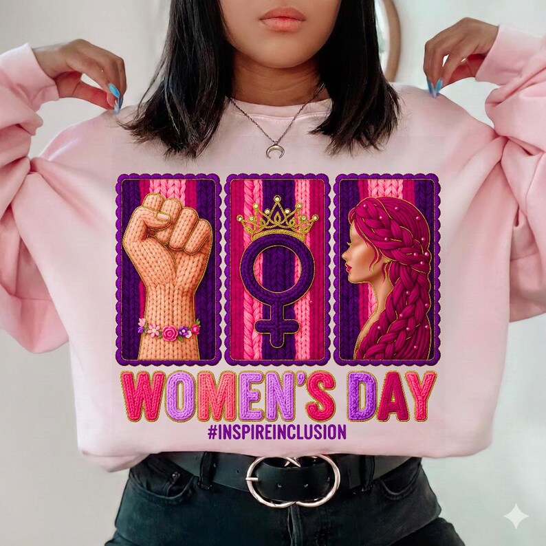 May include: Pink sweatshirt with a graphic design for Women's Day. The design includes a knitted fist, a female symbol with a crown, and a woman's profile. The text "WOMEN'S DAY" and "#INSPIREINCLUSION" are also featured.