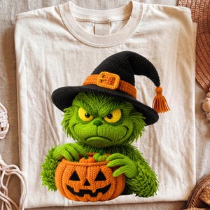 May include: A cream-colored t-shirt featuring a graphic of a green, fuzzy, crocheted-style creature wearing a black witch hat with an orange band and buckle. The creature is holding an orange crocheted pumpkin with a carved jack-o'-lantern face.