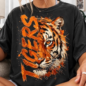 Puede incluir: Una camiseta de color negro carbón con un estampado gráfico de una cara de tigre en el lado derecho, en un estilo de pintura al óleo naranja y blanco. La palabra TIGERS está escrita verticalmente a la izquierda con una fuente de trazo grueso.