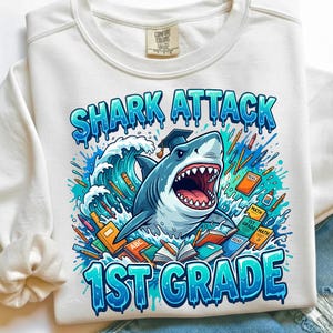 May include: A white crewneck sweatshirt featuring a graphic of a shark wearing a graduation cap emerging from a wave. The design includes school supplies like books, pencils, and crayons, with the text SHARK ATTACK 1ST GRADE in blue letters.