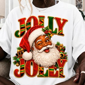 May include: A white t-shirt featuring a graphic of a winking Black Santa Claus wearing a red hat with holly. The text JOLLY JOLLY is printed in large red and green letters with a chevron pattern. Christmas ornaments and gifts surround the figure.