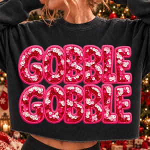 Puede incluir: Una sudadera negra de manga larga con la palabra GOBBLE GOBBLE en dos filas. Las letras están rellenas de lentejuelas rojas y rosas y delineadas con un borde rosa grueso. La sudadera tiene cuello redondo y un dobladillo corto.