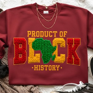 May include: A maroon sweatshirt with the text "PRODUCT OF BLACK HISTORY" in yellow and red, with a green Africa map in the center. A gold necklace is draped over the sweatshirt.