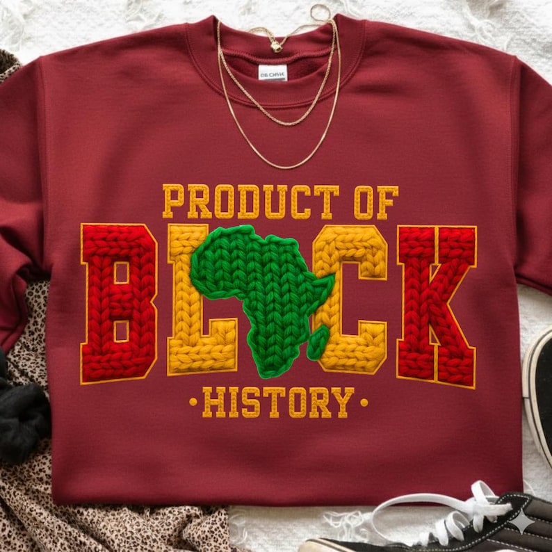 May include: A maroon sweatshirt with the text "PRODUCT OF BLACK HISTORY" in yellow and red, with a green Africa map in the center. A gold necklace is draped over the sweatshirt.