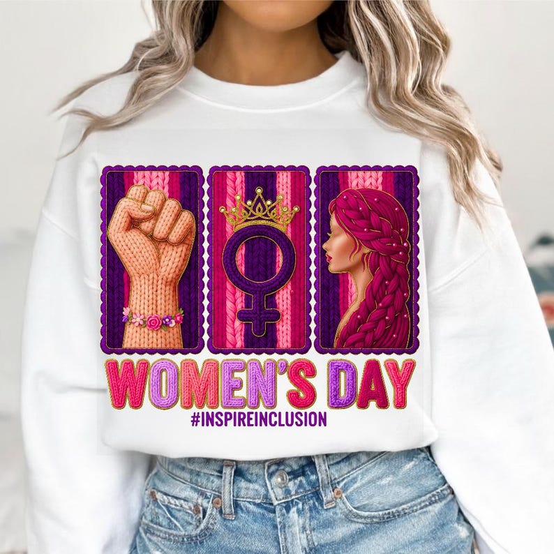 May include: White sweatshirt with a graphic celebrating Women's Day. The design includes a raised fist, a female symbol with a crown, and a woman's profile, all set against a purple and pink background. The text reads "WOMEN'S DAY" and "#INSPIREINCLUSION".