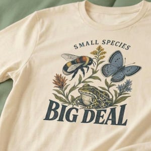 Può includere: Una t-shirt a maniche corte color crema con una stampa grafica raffigurante un bombo, una farfalla blu e una rana verde circondate da fiori di campo. Il testo "SMALL SPECIES" è stampato sopra l'illustrazione e "BIG DEAL" sotto di essa.
