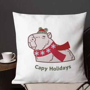 Puede incluir: Cojín cuadrado blanco con un capibara de dibujos animados que lleva una bufanda roja con diseños de copos de nieve y un gorro rojo y verde con pompones. El texto "Capy Holidays" está impreso debajo.