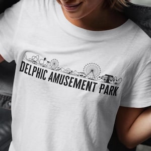 Peut inclure: T-shirt blanc avec un motif graphique noir représentant un horizon de parc d'attractions. Le texte "Delphic Amusement Park" est imprimé sous l'horizon.