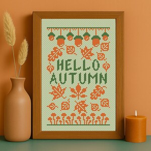 May include: A framed cross-stitch artwork with a wooden frame. The design features the words "HELLO AUTUMN" in green, surrounded by orange and green fall-themed motifs like acorns, leaves, and mushrooms. A vase with dried plants and a candle are in the foreground.
