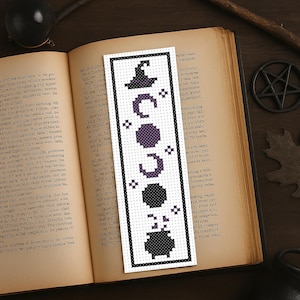 May include: A white bookmark with a black border, featuring a witch's hat, crescent moons, stars, a hexagon, and a cauldron in a cross-stitch design. The bookmark is placed on an open book with aged pages.