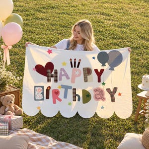 May include: A white rectangular blanket with scalloped edges featuring the text HAPPY BIRTHDAY in multi-colored, patterned, and solid letters. The design includes a red heart, three lit candles, and two gray balloons, accented with small colorful stars.