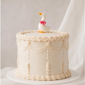 May include: A cylindrical cake with smooth off-white frosting, decorated with intricate piped swags, vertical floral motifs, and a ruffled border. A small white goose figurine with an orange beak and feet and a pink bow sits on top.