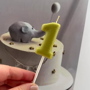 May include: A lime green number one shaped candle attached to a white stick. The candle features a textured, slightly puffed appearance with a single thin wick protruding from the top. The design is simple, solid in colour, and functional.