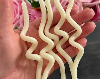 May include: Four off-white taper candles with a distinct wavy, serpentine shape. Each candle features a smooth wax finish and a single cotton wick at the top. The candles are held together in a cluster, showcasing their elongated, curved design.