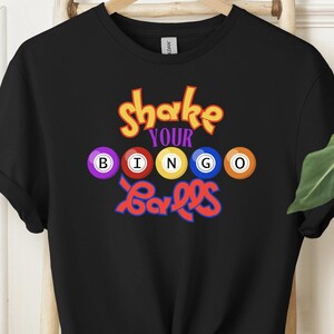 May include: Black t-shirt with the text "Shake Your Bingo Balls" in colorful letters. The text is surrounded by five colorful bingo balls with numbers.