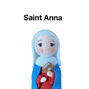 May include: Crocheted doll of Saint Anna, wearing a blue robe over a red dress, holding a book in her hands. The doll has a white head scarf and a painted face.