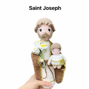 May include: Crocheted amigurumi doll of Saint Joseph holding a small crocheted baby Jesus doll. The dolls are beige and brown with white accents. Saint Joseph is holding a bouquet of white flowers.