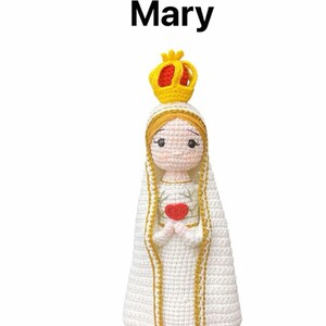 May include: Crocheted doll of Mary, the mother of Jesus, wearing a white robe with gold trim and a crown. She is holding a red heart in her hands.