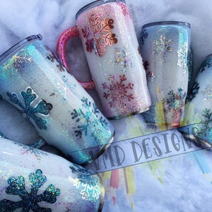 May include: Set of five stainless steel tumblers with a white base and snowflake designs. The tumblers have a glittery finish in shades of blue, pink, and white. The tumblers have a handle and are perfect for hot or cold beverages.