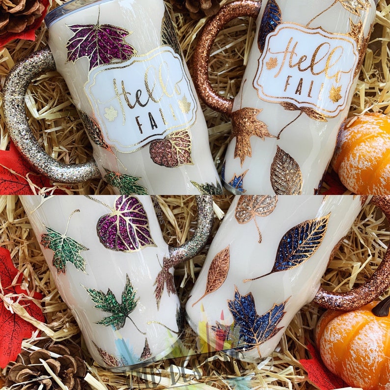 May include: Three white tumblers with handles, each decorated with a different color glitter and leaf design. The tumblers have the words "Hello Fall" written on them.