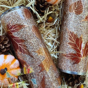 May include: Two gold glitter tumblers with a fall leaf design. The leaves are a deep red and brown color. The tumblers are sitting on a bed of straw.