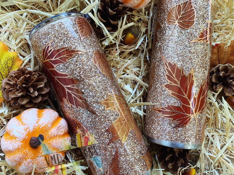 May include: Two gold glitter tumblers with a fall leaf design. The leaves are a deep red and brown color. The tumblers are sitting on a bed of straw.