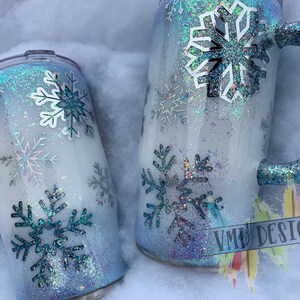 May include: Two white and blue glitter tumblers with snowflake designs. The tumblers have blue glitter handles. The larger tumbler has a white snowflake design and the smaller tumbler has a blue snowflake design. The tumblers are sitting on a white surface. VMD Designs is written on the bottom right corner of the image.