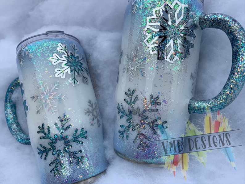 May include: Two white and blue glitter tumblers with snowflake designs. The tumblers have blue glitter handles. The larger tumbler has a white snowflake design and the smaller tumbler has a blue snowflake design. The tumblers are sitting on a white surface. VMD Designs is written on the bottom right corner of the image.