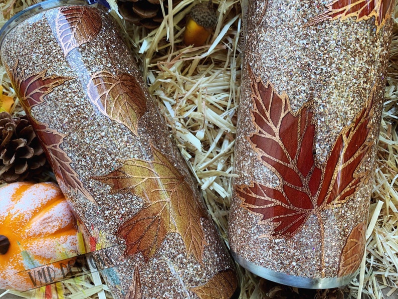 May include: Two gold glitter tumblers with a fall leaf design. The leaves are in shades of brown, gold, and red. The tumblers are sitting on a bed of straw.