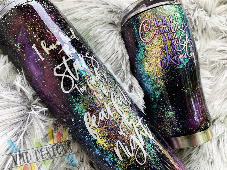 May include: Two stainless steel tumblers with a galaxy-inspired design. The larger tumbler has the text "Have Courage and Be Kind" on it. The smaller tumbler has the text "I have heard the stars too faintly to be fearful of the night".