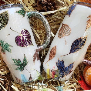 May include: Two white tumblers with handles, decorated with fall leaves in various colors and glitter. The leaves are gold, green, purple, and blue.