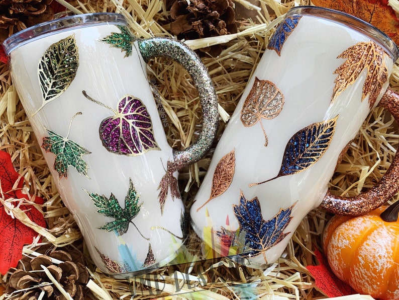 May include: Two white tumblers with handles, decorated with fall leaves in various colors and glitter. The leaves are gold, green, purple, and blue.