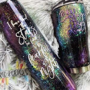 May include: Two stainless steel tumblers with a galaxy-inspired design. The larger tumbler has the text "I have loved the stars too fondly to be fearful of the night". The smaller tumbler has the text "Have courage and be kind".